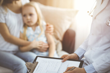 Doctor and patient. Pediatrician using clipboard while examining little girl with her mother at home. Sick and unhappy child at medical exam - 668841978