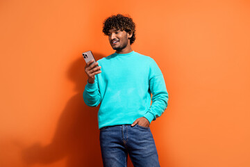 Photo of young indian happy guy with curly hair wearing turquoise pullover using smartphone dating app isolated on orange color background