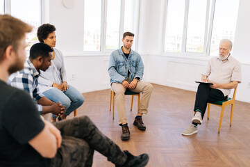 Portrait of frustrated Caucasian young man drinker talking about alcohol addiction abuse sitting in circle, speak to diverse addicted people at group therapy session. Concept of mental health.