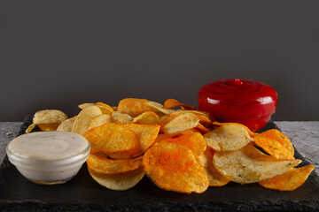 Side view of heap of potato chips with two dipping sauces on black slate serving board on grey plaster table. Flavorful Dip Pairings