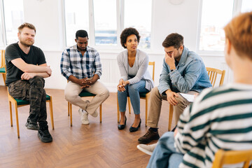 Front view of multi-ethnic different ages people having discussion about addiction at group therapy sitting in circle. Men and women talking about mental health problems with psychiatrist at meeting.