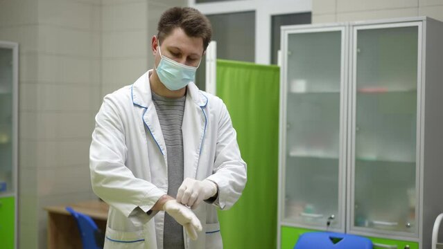 A male medical worker, wearing a white coat and a mask, puts on medical pants and prepares for work