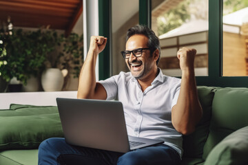 Handsome middle age arab man smiling and rejoicing after success. Happy man celebrating business success on sofa in living room with computer.
