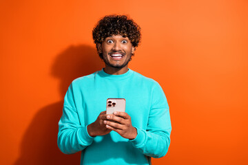 Photo of young surprised funny positive man indian tik tok user wearing teal pullover using smartphone isolated on orange color background - obrazy, fototapety, plakaty