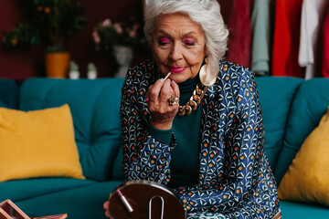 Stylish senior woman in fashionable wear applying lipstick while sitting on the couch at home