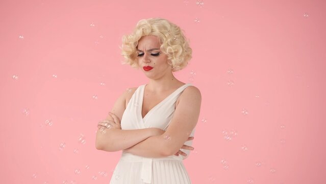 Portrait of a disgruntled woman in the image of Marilyn Monroe surrounded by soap bubbles in studio on pink background. Offended woman standing with her arms crossed on her chest.