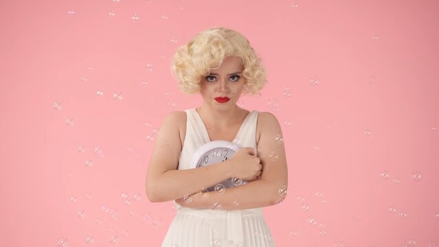 Sad woman holding round clock, covering dial with hands. Woman in image on Marilyn Monroe, in studio surrounded by soap bubbles on pink background. Concept of time.