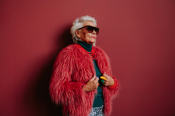 Fashionable elderly woman in stylish eyeglasses and trendy wear standing on red background