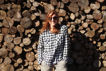 Mulher sorridente de óculos de sol e camisa quadriculada em frente a pilhas de madeira