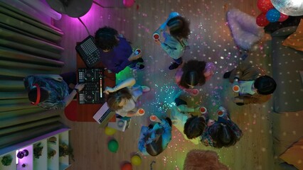 Group of teenagers dancing in a decorated party room, holding cups. A man is mixing music on a dj console. Neon colorful lights. Top view.