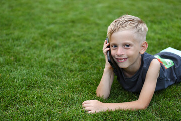 Naklejka na ściany i meble A cute little child, blond, lies on the green grass and talks on the phone.