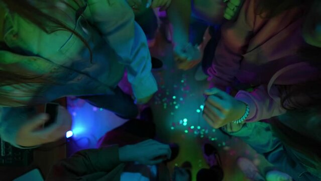Group of teenagers dancing in a decorated party room with a disco ball. A man is mixing music on a dj console. Neon colorful lights. Top view, zoom out.