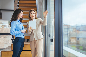 Two business women having a discussion, they're standing in an office and using a tablet. Professional women making a to do list as part of their project planning.