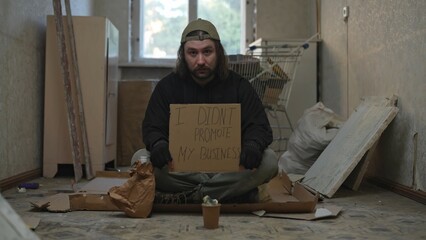 Homeless poor man sitting in a room of an abandoned building. He is holding a piece of cardboard,...
