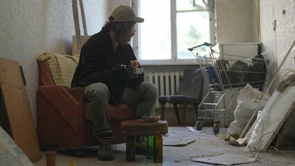Homeless poor man sitting in a room of an abandoned building. He eats some food out of disposable...