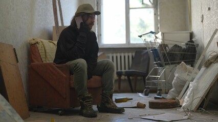 Homeless poor man sitting in a room of an abandoned building filled with his meager belongings. He...