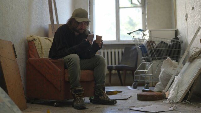 Homeless poor man sitting in a room of an abandoned building filled with his meager belongings. He is holding a smartphone and a cup in his hands.