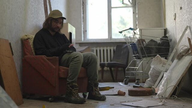Homeless poor man sitting in a room of an abandoned building filled with his meager belongings. He takes a few money bills, notes out of his pocket and starts counting his savings.