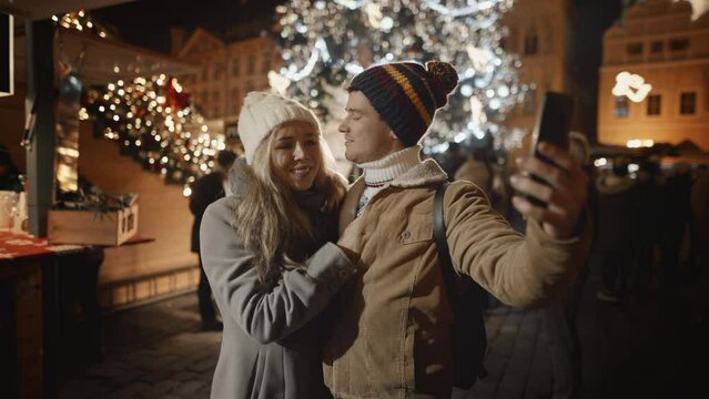 Caucasian couple or family kiss take selfie at Christmas New Year holidays at illuminated background of decorated town square. Woman and man in love enjoy Eve winter vacation. Celebration of Christmas