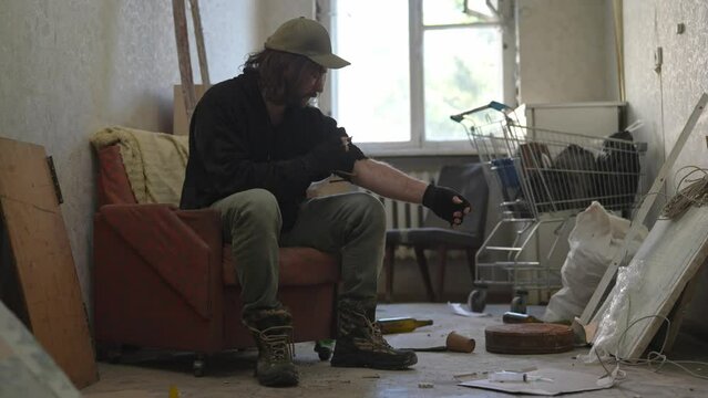 Homeless poor man sitting in a room of an abandoned building filled with his meager belongings. He tightens the tourniquet on his arm and injects an illegal substance, drug into his vein.