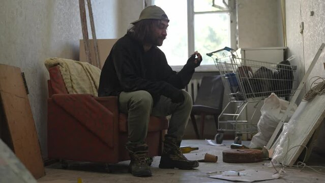 Homeless poor man sitting in a room of an abandoned building filled with his meager belongings. He lights a cigarette or a joint, blunt and starts smoking it.