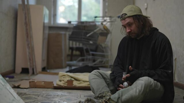 Homeless poor man sitting in a room of an abandoned building filled with his meager belongings. He drinks alcohol from a bottle, looking frustrated.