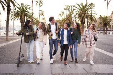 Diverse multicultural group young millennial friends walking along urban street palm trees. University people happy strolling outside on way to campus. Concept of cheerful students together.  - obrazy, fototapety, plakaty