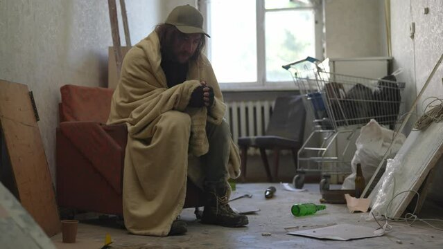 Homeless poor man covered in a blanket sitting in a room of an abandoned building filled with his meager belongings. He takes pills from a bottle.