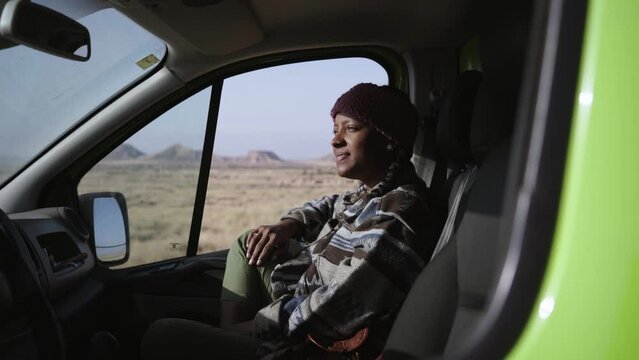 Happy young African American woman enjoys looking smiling at scenery inside caravan. Black girl co-driver rests relaxed with background bardenas desert. Generation z road trips on vacation in nature.