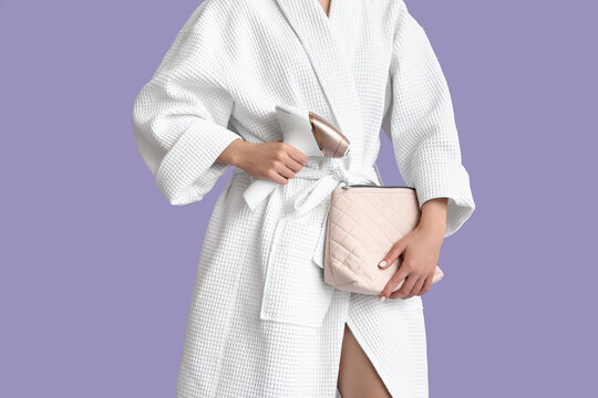 Beautiful young woman in bathrobe with cosmetic bag and modern photoepilator on lilac background