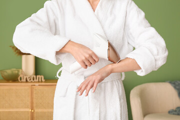 Beautiful young woman in bathrobe epilating her arm with modern photoepilator at home