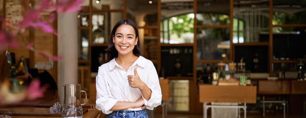 Confident asian businesswoman, showing thumbs up, standing near entrance of her cafe or restaurant,...