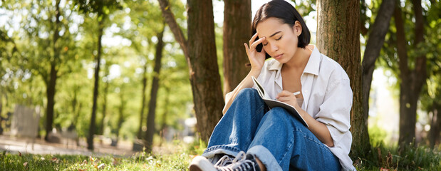 Portrait of sad asian girl writing in her diary and feeling uneasy, sitting in park alone under tree, expressing her distress in notebook