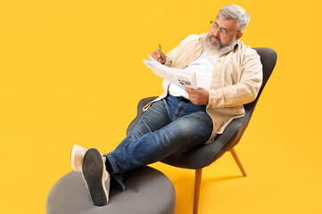 Fototapeta premium Mature man with pen and newspaper sitting on armchair against yellow background