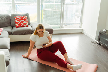Cute young plus size woman sitting on yoga mat, holding mobile phone, text message