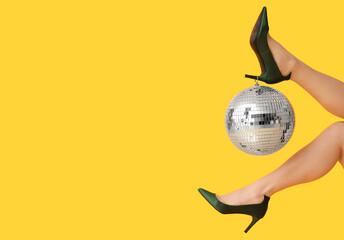 Female legs in stylish high heels with disco ball on yellow background