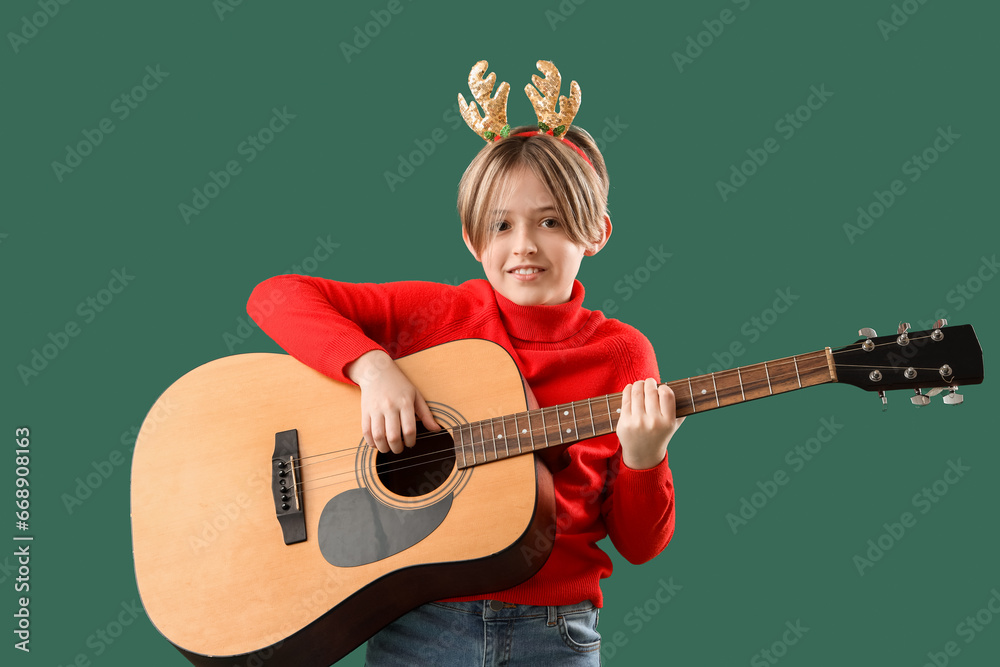 Sticker Cute little boy in Christmas deer horns playing guitar on green background