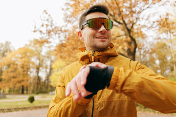 Portrait of a man Motivation for an active life. A running coach uses a smart watch and a fitness app on an autumn day. In sports glasses for running, smiling. - obrazy, fototapety, plakaty