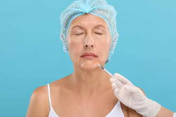 Doctor giving lips injection to senior woman on light blue background. Cosmetic surgery