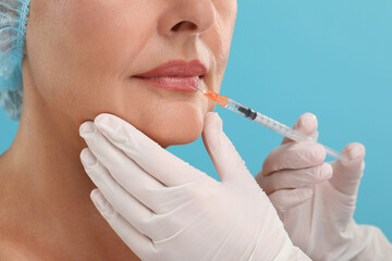 Doctor giving lips injection to senior woman on light blue background, closeup. Cosmetic surgery
