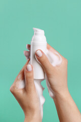 Woman with foam on hands holding bottle of skin cleanser against green background, closeup. Cosmetic product