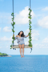 Happy young asian woman relaxing on a swing on summer day outdoors  on with Andaman blue sea and sky background. Summer Vacations lifestyle. Phuket, Thailand.
