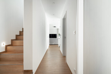 Entrance hall in family house. On left side there is staircase to upper floor. Corridor with several doors leads to other rooms and ends with living room. Racks are white, floor is wooden.