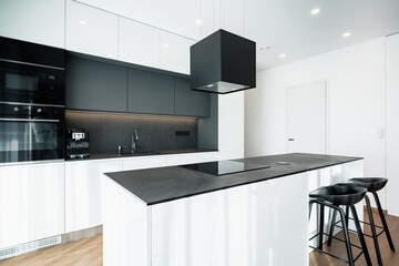 Modern kitchen with island.It has clean lines,black and white design and appliances.There is also electric oven,microwave,coffee machine,sink with tap,hood and hob.There are two bar stools at island.
