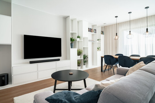 Modern living room with dining area in clean design with builtin furniture and window.There're large TV with sound bar,coffee table,many decorative plants,massive table with chairs and ceiling lights.