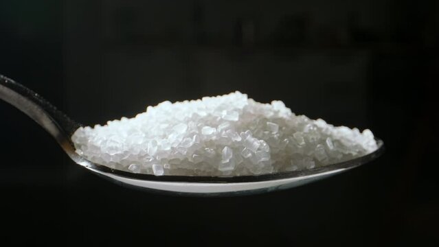 Spoon of white sugar. Close up slow motion black background. Insidious nature of sugar allows it to permeate foods we least suspect making it imperative to be mindful of hidden sugar content. Diabetes