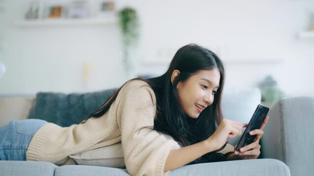 Happy young asian woman relaxing at home. Female smile lying on sofa and holding mobile smartphone. Girl using video call to friend