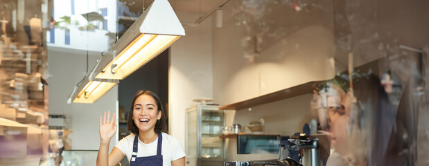 Happy asian girl barista works in coffee shop, waves at client, processing orders behind counter in...