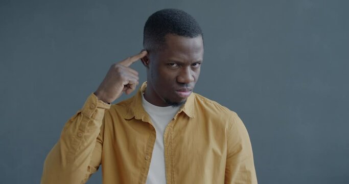 Slow motion portrait of African American man frowning and making Are you Crazy hand gesture twisting finger at temple on grey color background
