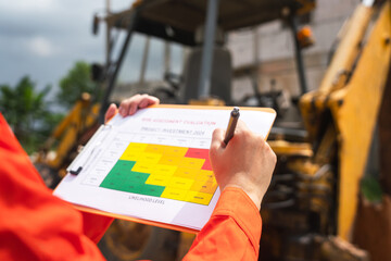 The operation supervisor is using ballpoint pen to marking on the risk assessment matrix form, to inspection the backhoe machine that using at construction worksite. - obrazy, fototapety, plakaty